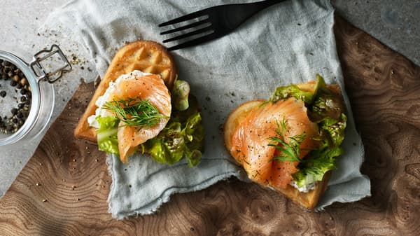 Two heart shaped VÅFFLOR waffles with salmon, cream cheese, dill and lettuce on a piece of fabric next to a fork.