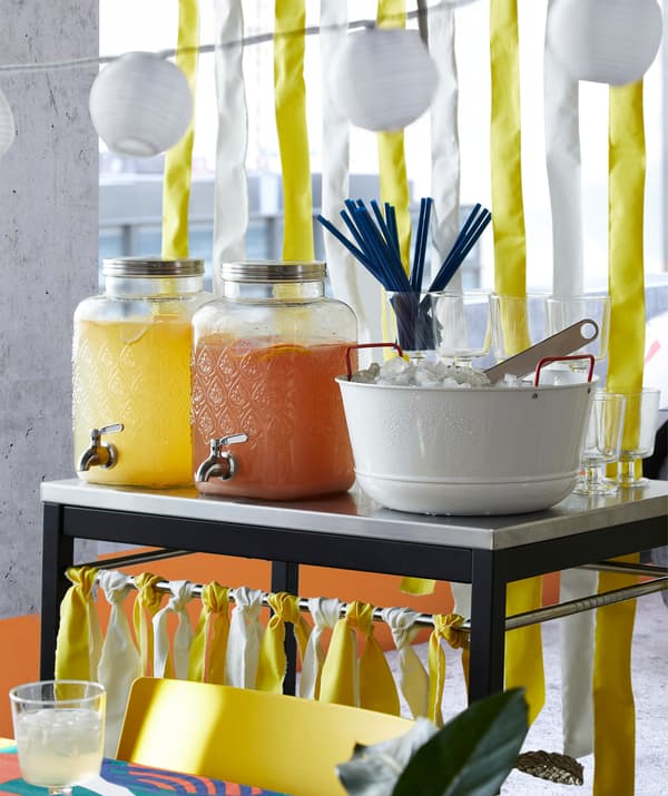 Two IKEA FÖRFRISKNING glass jars with taps in stainless steel and filled with yellow and orange fruit juices for a party.