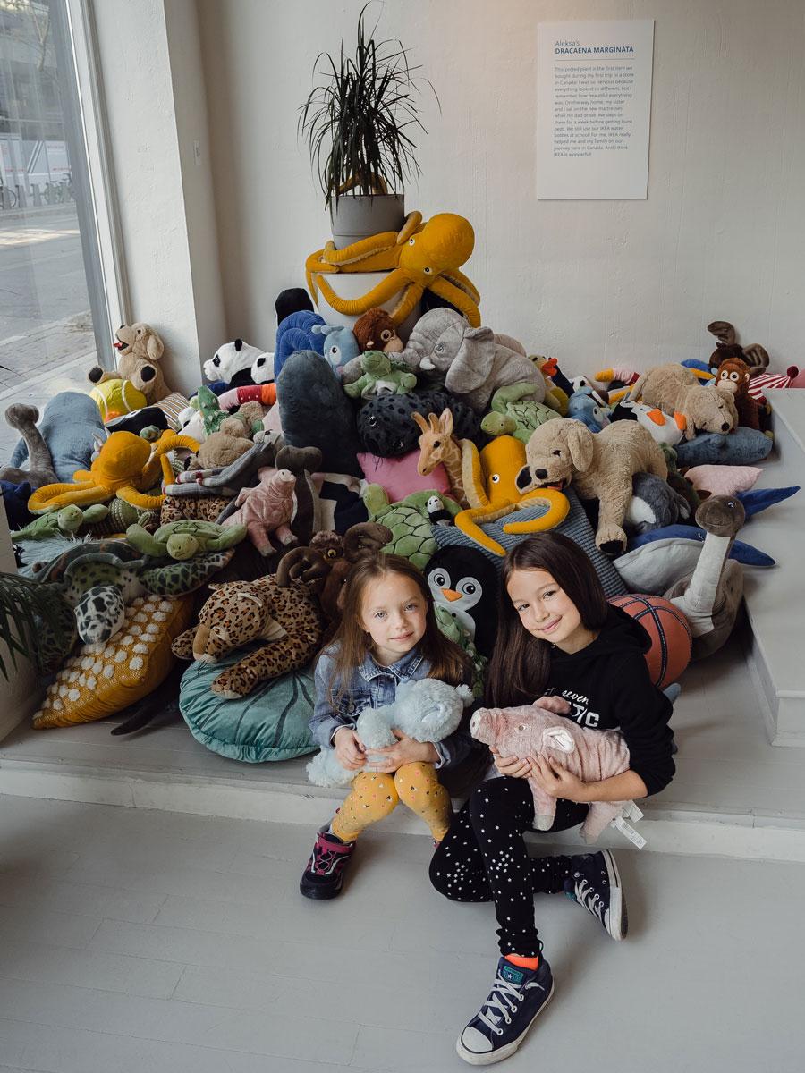 Two kids sit in front of a display showing a large number of soft toys and a plant placed in the center of the display.