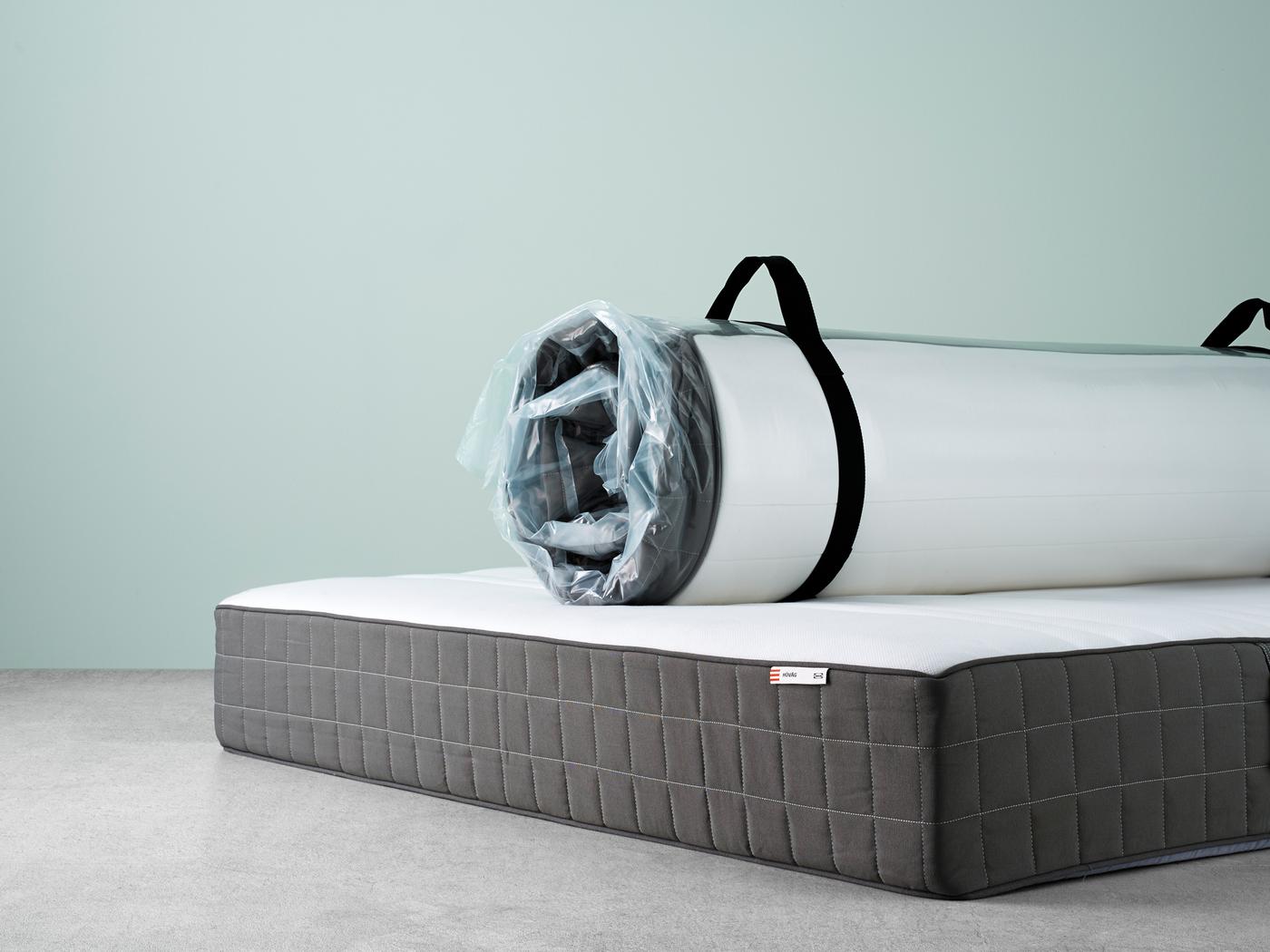 Two mattresses with no sheets on them. One is opened flat on the floor, the other is rolled up in packaging on top.