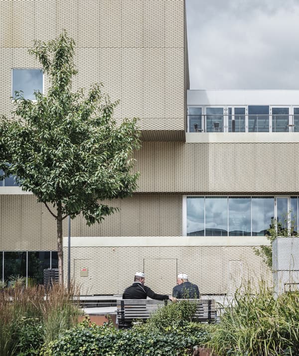 Two men sit on a bench in a garden outside a building covered in golden metal.