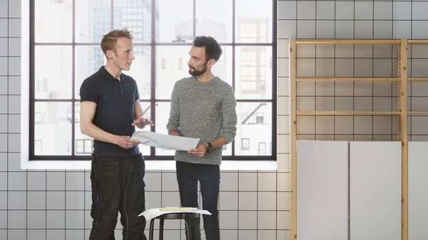Two men standing in front of a wall with square tiling and shelving, talking to each other while holding documents.