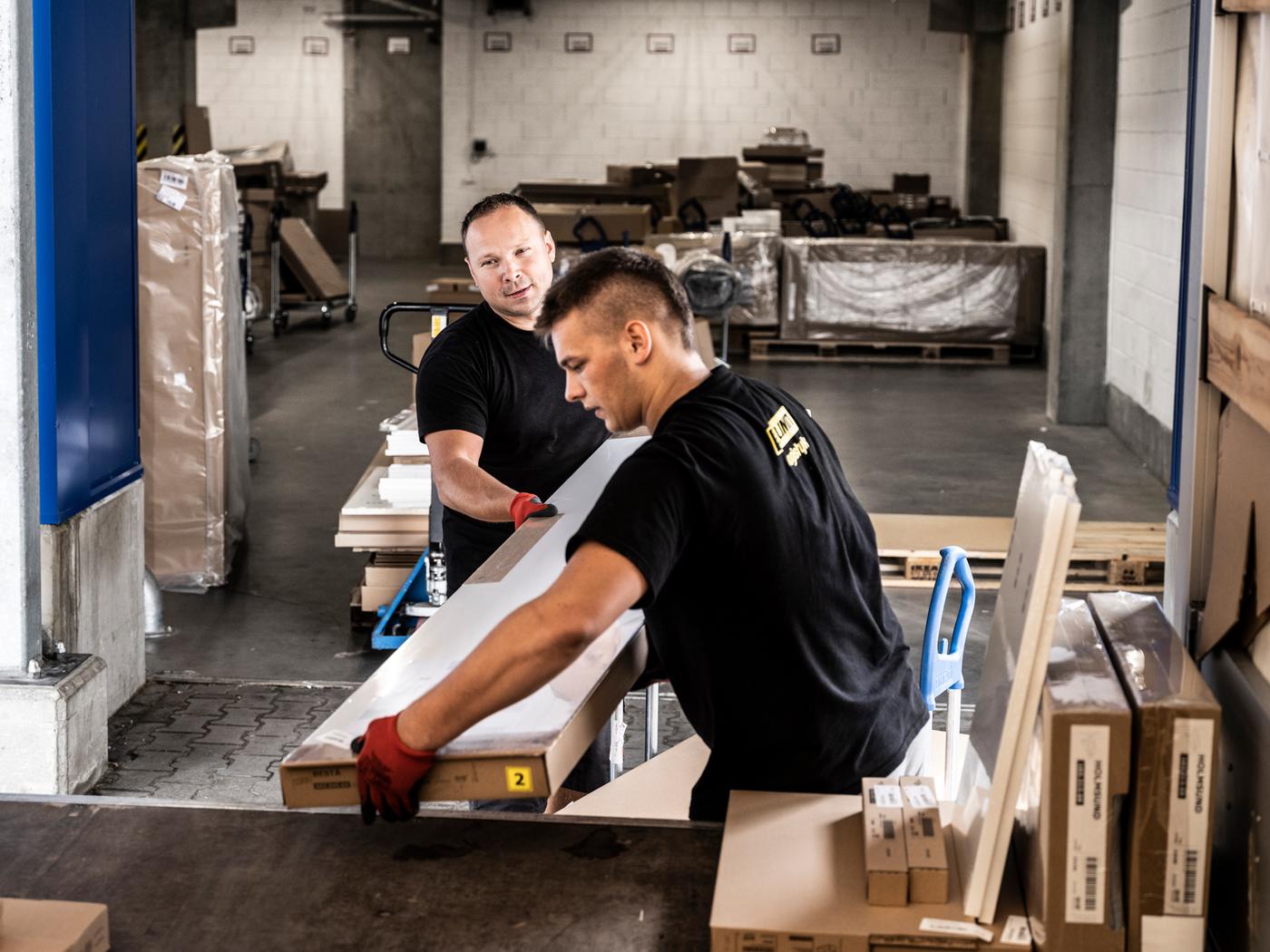 Two men wearing black t-shirts and red work gloves loading IKEA flatpacks from a trolley into the back of a truck.