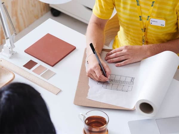 Two people at a table, one drawing a floor plan on paper while the other watches, with two cups of tea, a laptop, color swatches, a ruler, and documents lying on the table, among other things.