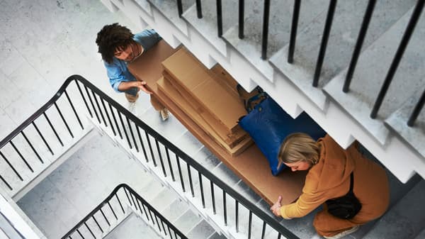 Two people carry several large IKEA flat-pack boxes and a blue IKEA bag up a flight of grey stairs in an apartment building.