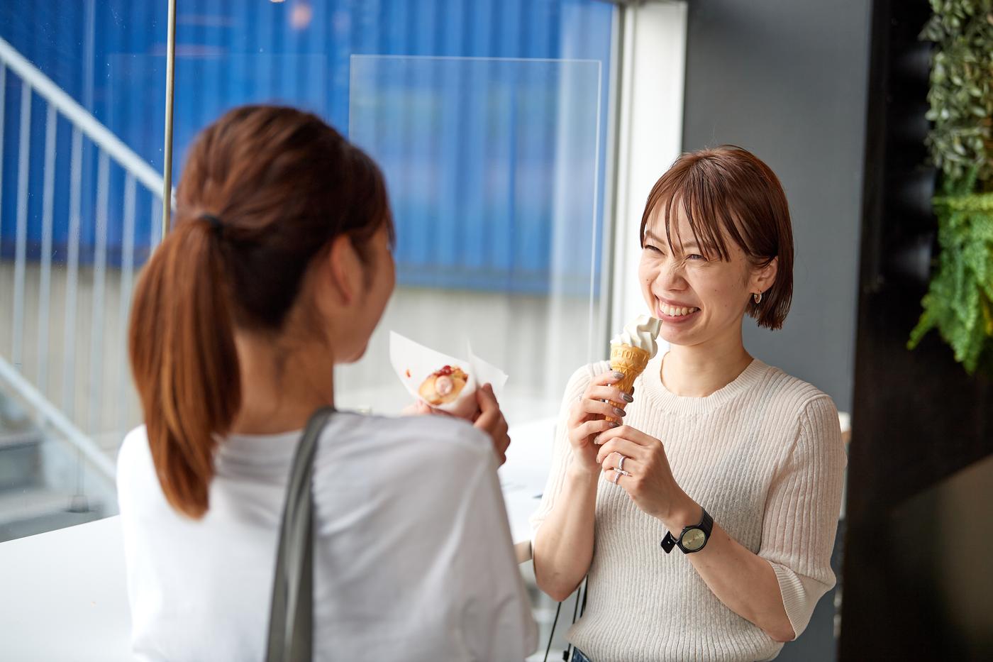 Two people enjoying ice cream by a window; one faces the camera, the other stands with their back turned.