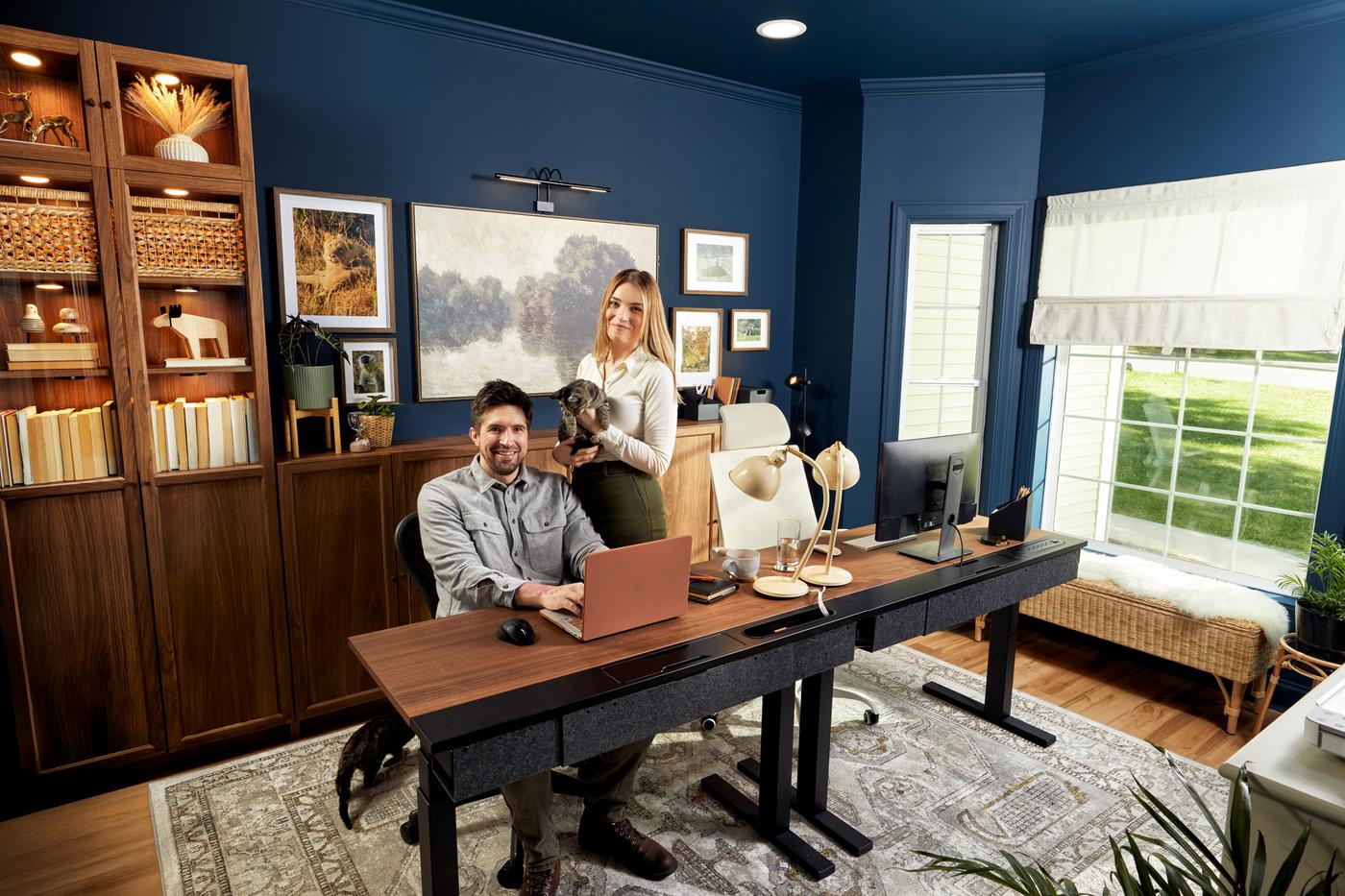 Two people in a blue office room with a desk and monitors.