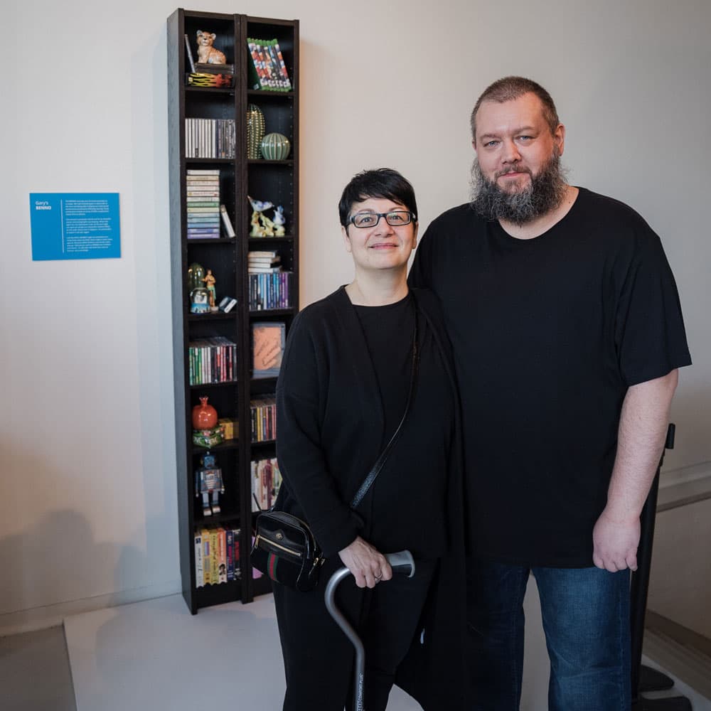 Two people pose in from of an IKEA BENNO bookcase, holding different items such as decorations, books, toys, and gaming CDs.