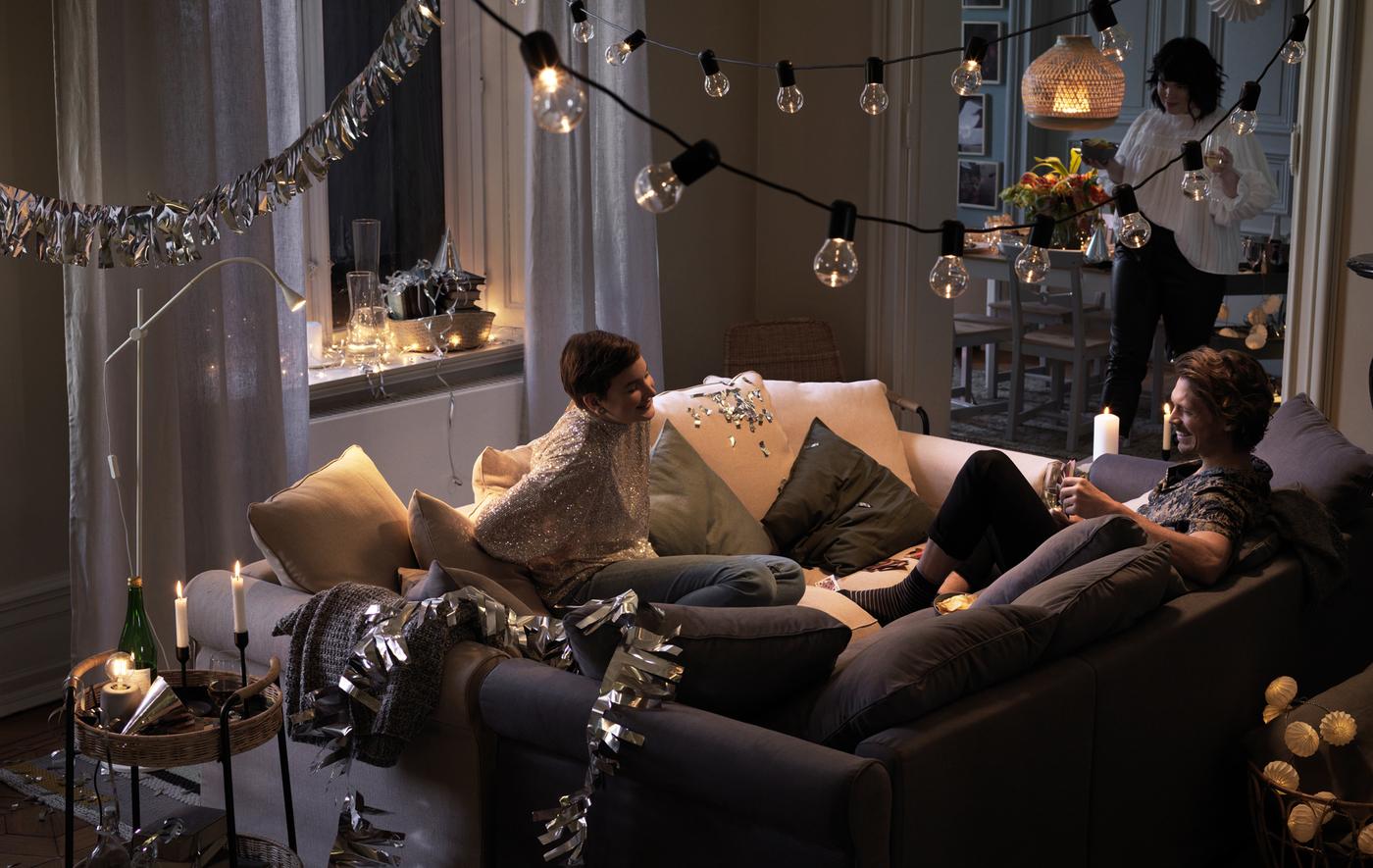 Two people sit together in two sofas that have been pushed together in the middle of a room, laughing and chatting.