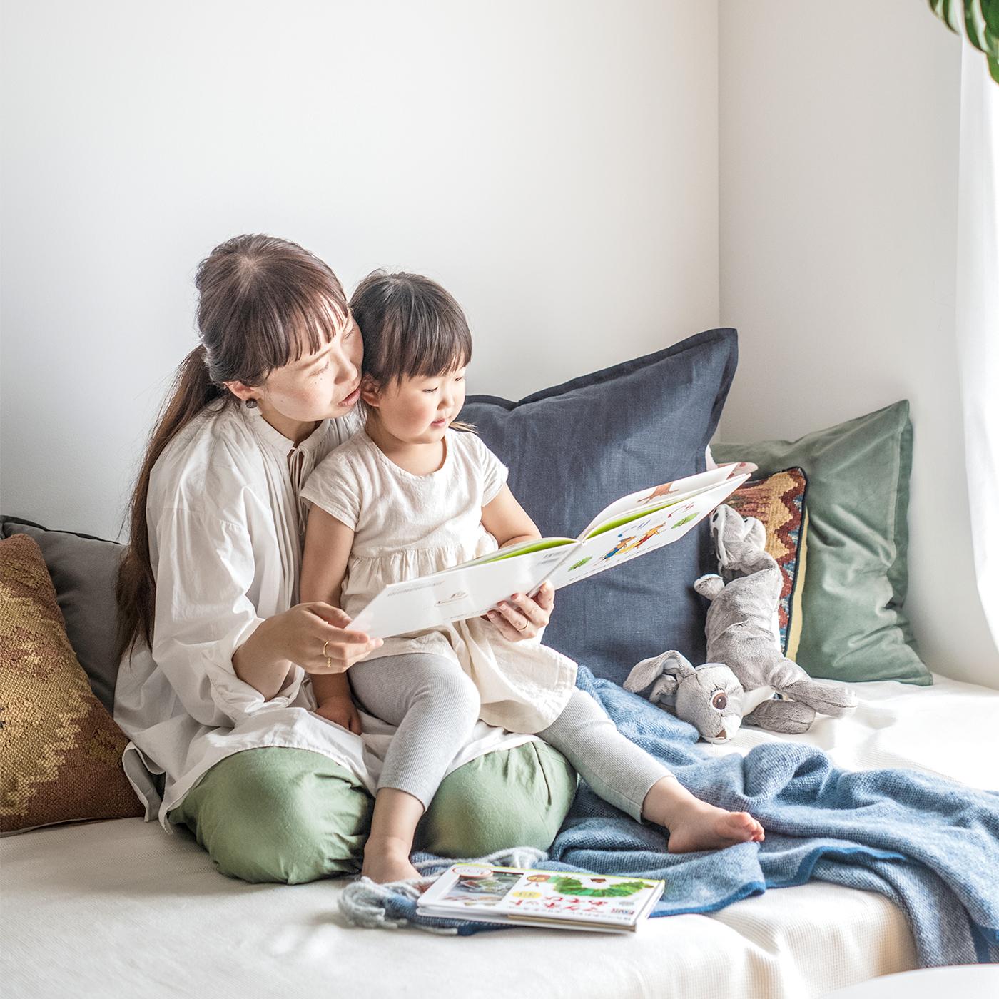 Two people sitting on a sofa reading a book together. Cushions and a stuffed animal are visible in the background.