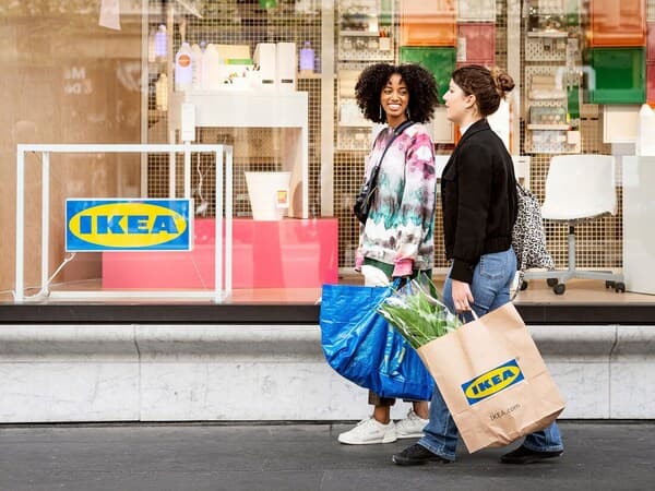 Two people who have been shopping at IKEA, one carrying a blue FRAKTA bag and the other an IKEA paper bag containing plants.