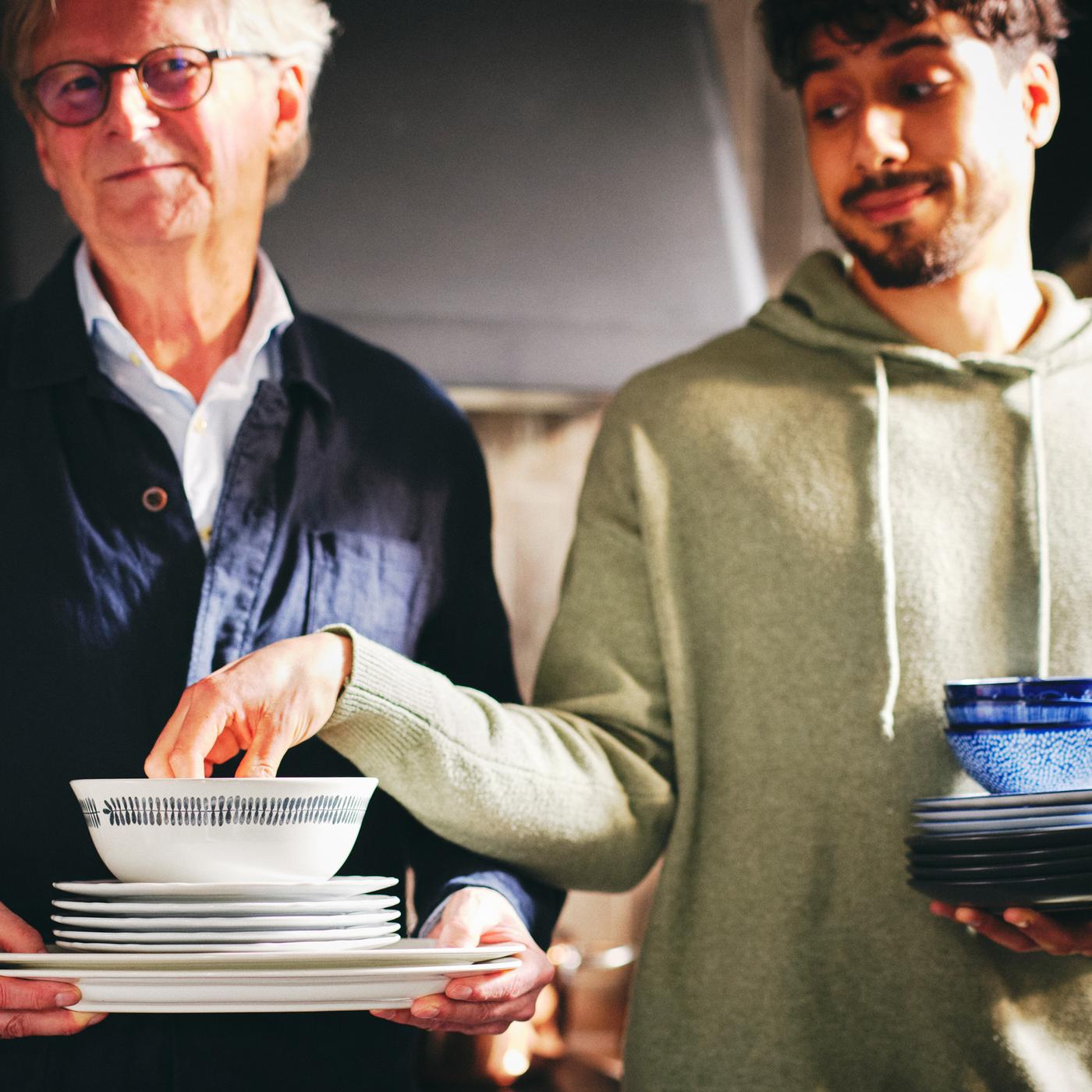 Two people with one of them holding a stack of white UPPLAGA serving and side plates and a white/patterned FRIKOSTIG bowl.