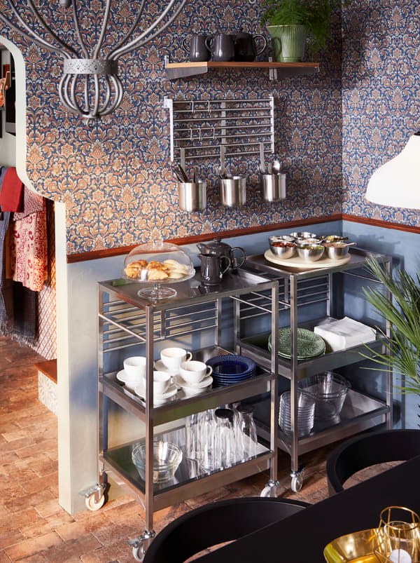 Two stainless steel KUNGSFORS trolleys holding glasses, bowls, cups, saucers and food, against a wall, pots of cutlery hang above.