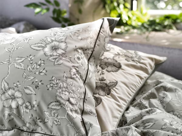 Two versions of IKEA PRAKTBRÄCKA pillowcases in white and grey cotton with vintage flower designs lying on a bed.