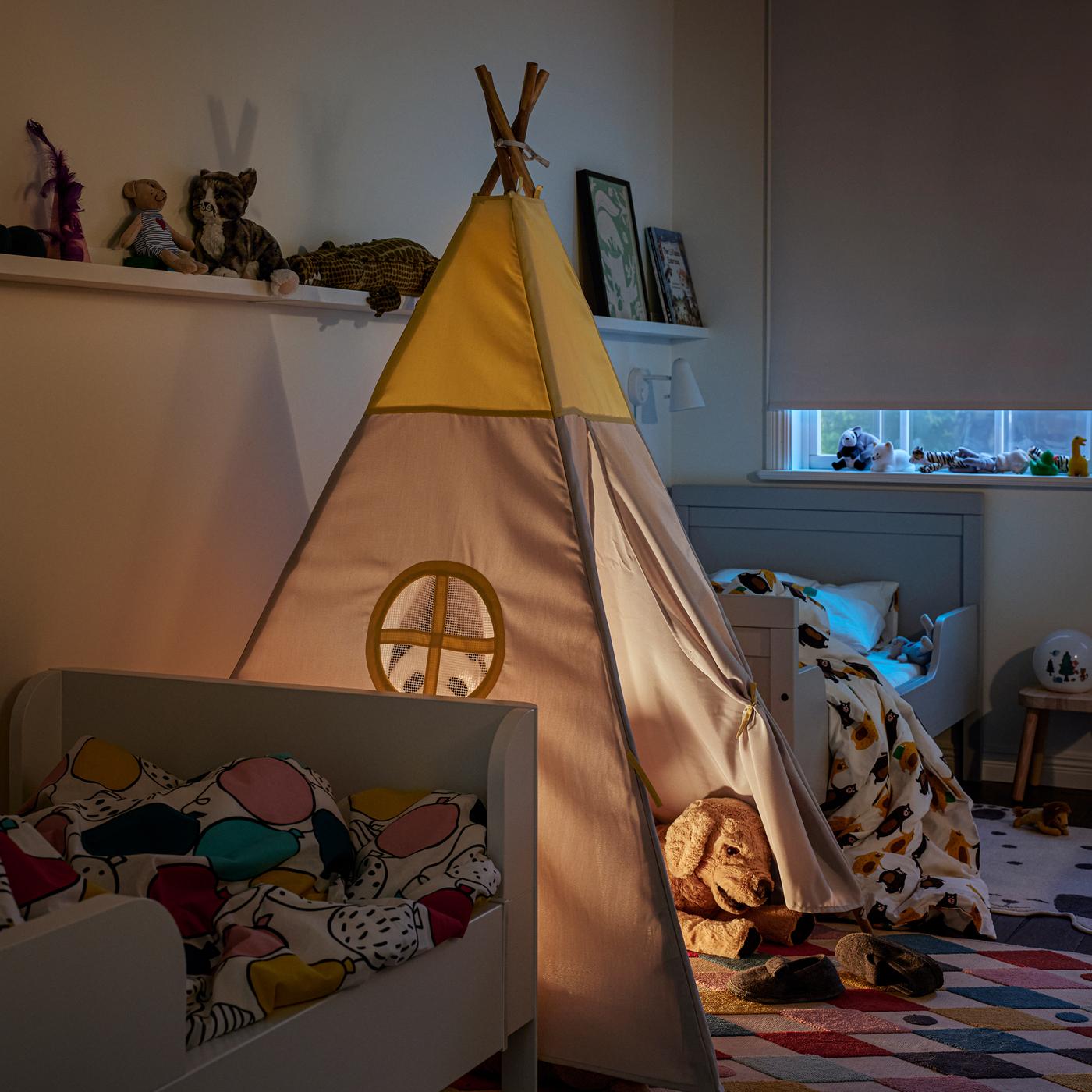 Two white extendable beds, a HÖVLIG children’s tent on a multicolour rug and a partially drawn white block-out roller blind.