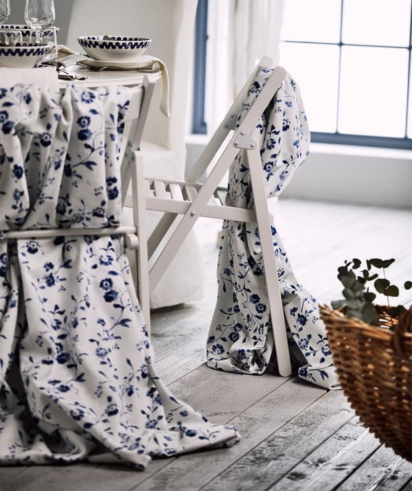 Two white folding chairs draped with white and blue flower patterned BLÅGRAN fabric, standing next to a festive table.