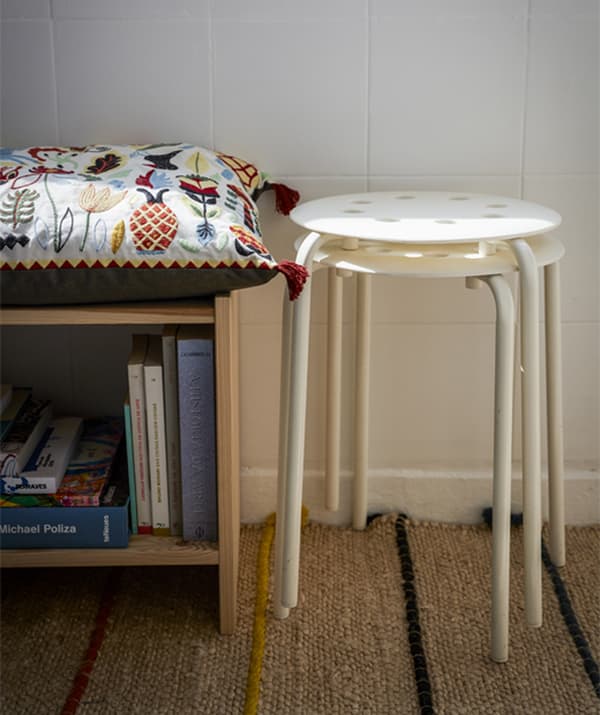 Two white stools stacked up next to a low bookshelf with a seat cushion on top.