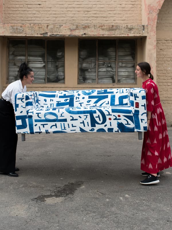 Two women holding either end of a sofa with a blue and white textile design on a tarmac surface outside a brick storehouse.
