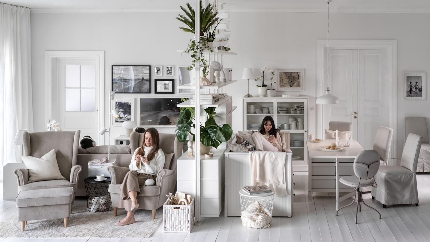 Two women seated, one in a beige STRANDMON armchair, on either side of an ELVARLI storage unit in a large, light living room.