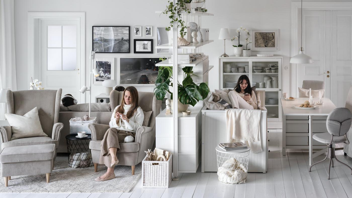 Two women seated, one in a beige STRANDMON armchair, on either side of an ELVARLI storage unit in a large, light living room.