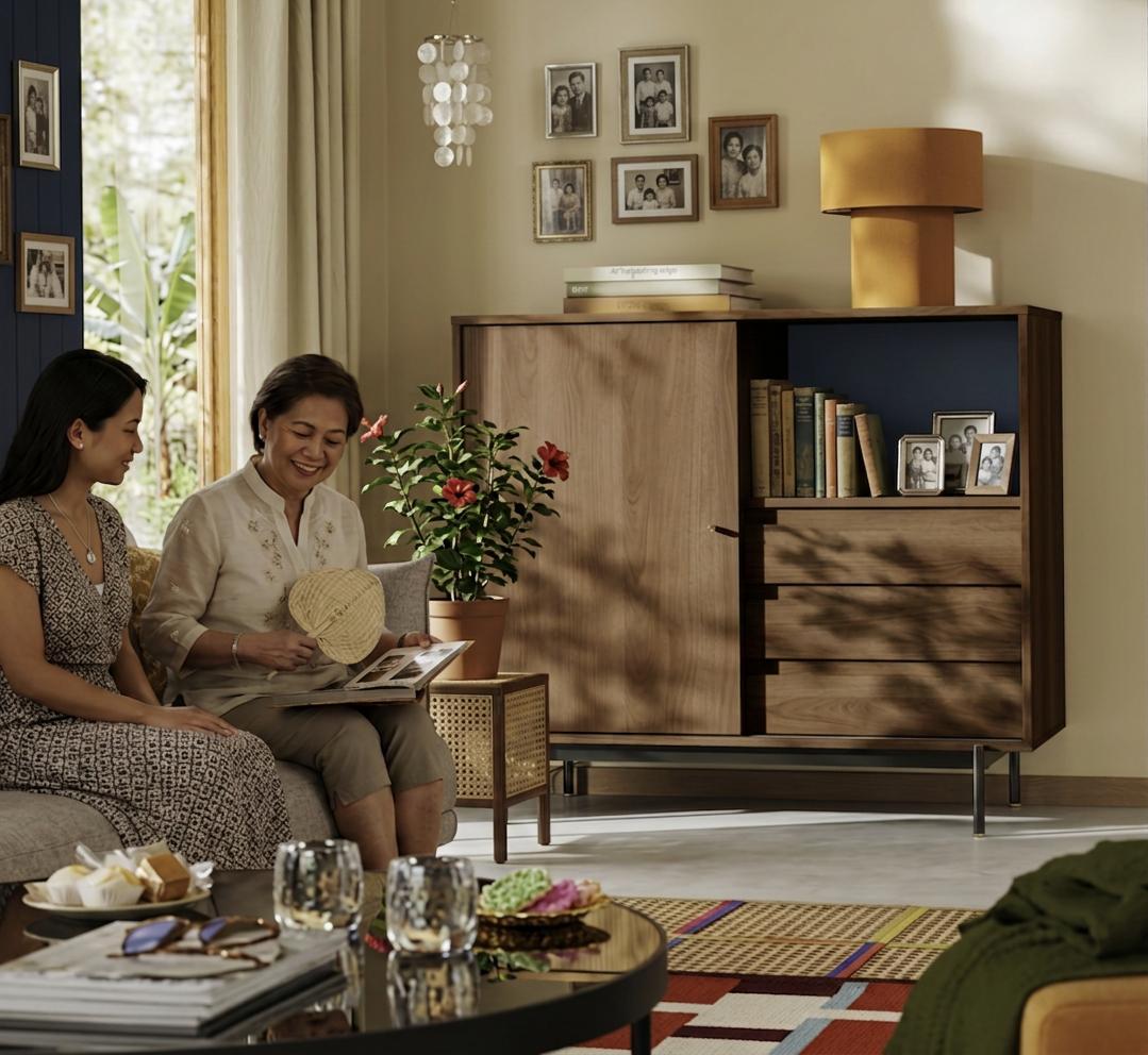 Two women sit together on a sofa, smiling as they look through a photo album in a warm, sunlit living room with vintage decor.