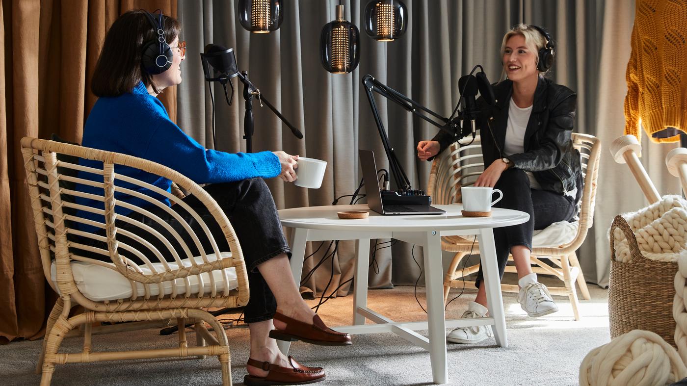 Two women wearing recording equipment sit in BUSKBO rattan armchairs, facing each over a white KRAGSTA coffee table.