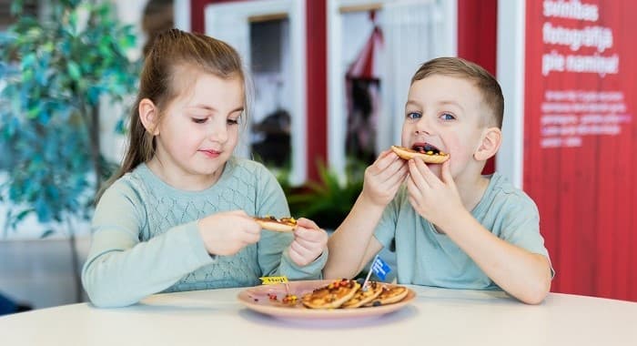 Two young children sitting at a table enjoying small pancakes topped with jam and sprinkles, with a plate full of pancakes between them. The background shows a red wall and a cozy, indoor setting.