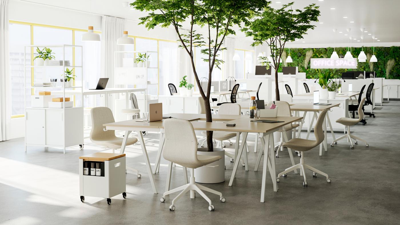Ufficio open space con diversi gruppi di scrivanie e sedie in beige/bianco, con piante verdi nel mezzo.