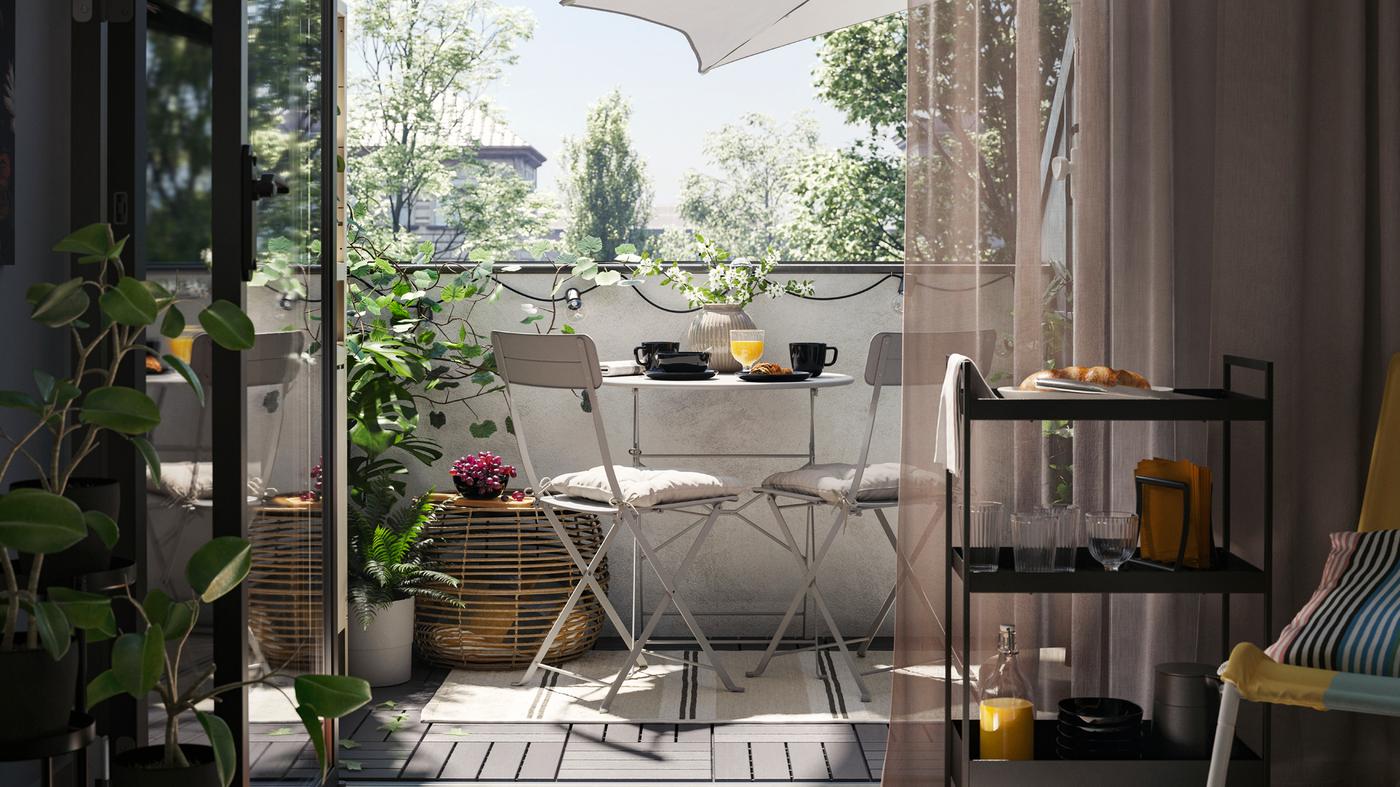 Un balcone con un tavolo e due sedie da giardino SUNDSÖ in grigio su un tappeto, oltre a un tavolino, piante e un ombrellone - IKEA.