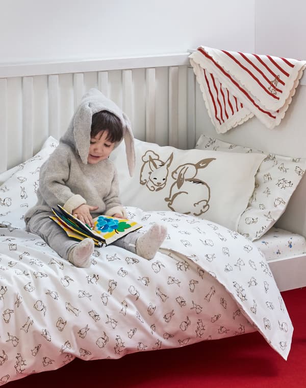 Un bambino che indossa una tutina con le orecchie da coniglio è seduto in un lettino con copripiumino e federa RÖDHAKE, mentre sfoglia un libro per bambini - IKEA