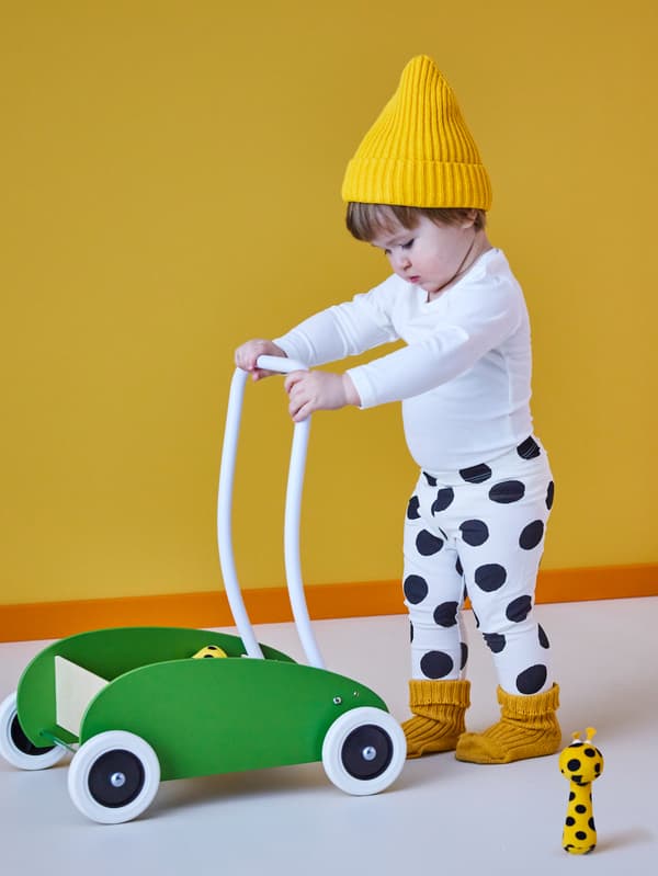 Un bambino con un cappello di lana giallo spinge un carrellino primi passi MULA verde, accanto a lui un morbido peluche e sullo sfondo una parete gialla - IKEA