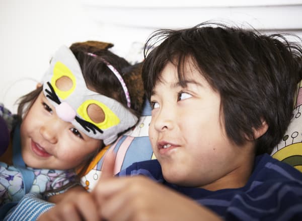 Un bambino e una bambina con una maschera da gatto sollevata sulla fronte, seduti sul divano a parlare – IKEA