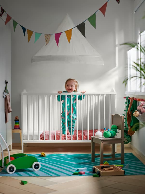 Un bambino in piedi in un lettino SMÅGÖRA bianco e mobili per bambini e giocattoli nei toni del rosso, verde e azzurro.