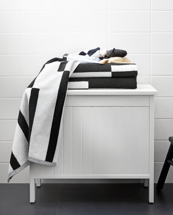 Un banc avec rangement SILVERÅN blanc surmonté de serviettes au motif graphique noir et blanc, dans une salle de bain.