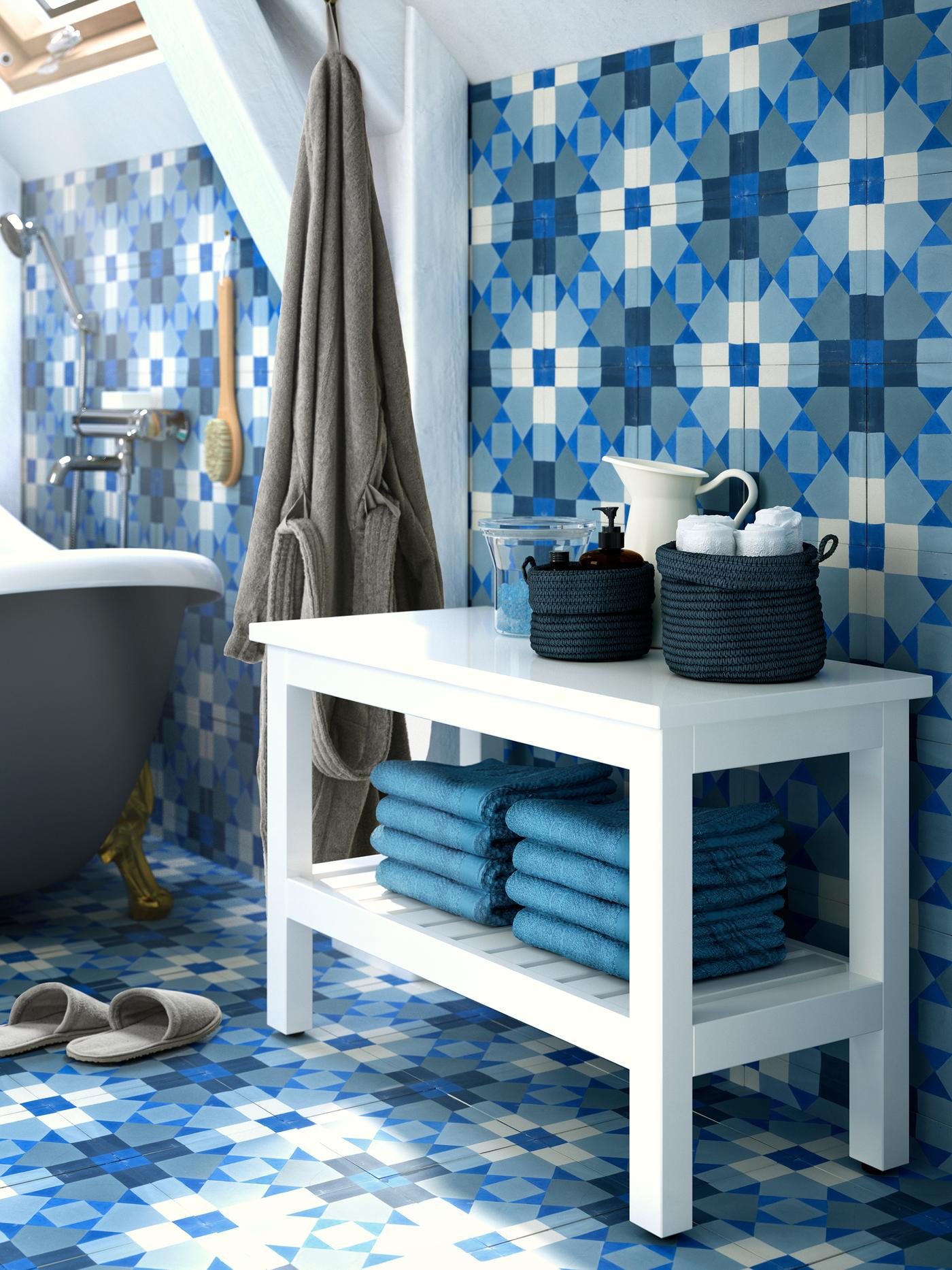  Un banco HEMNES blanco con toallas en un estante inferior y cestas con accesorios en la parte superior, con paredes y suelo de azulejos azules.