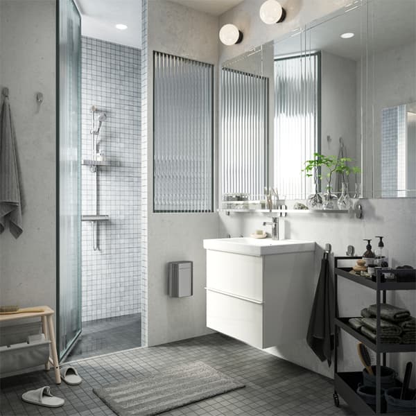 Un baño blanco con gris con una regadera, un lavabo blanco y un carrito negro para almacenar toallas, jabones y más.