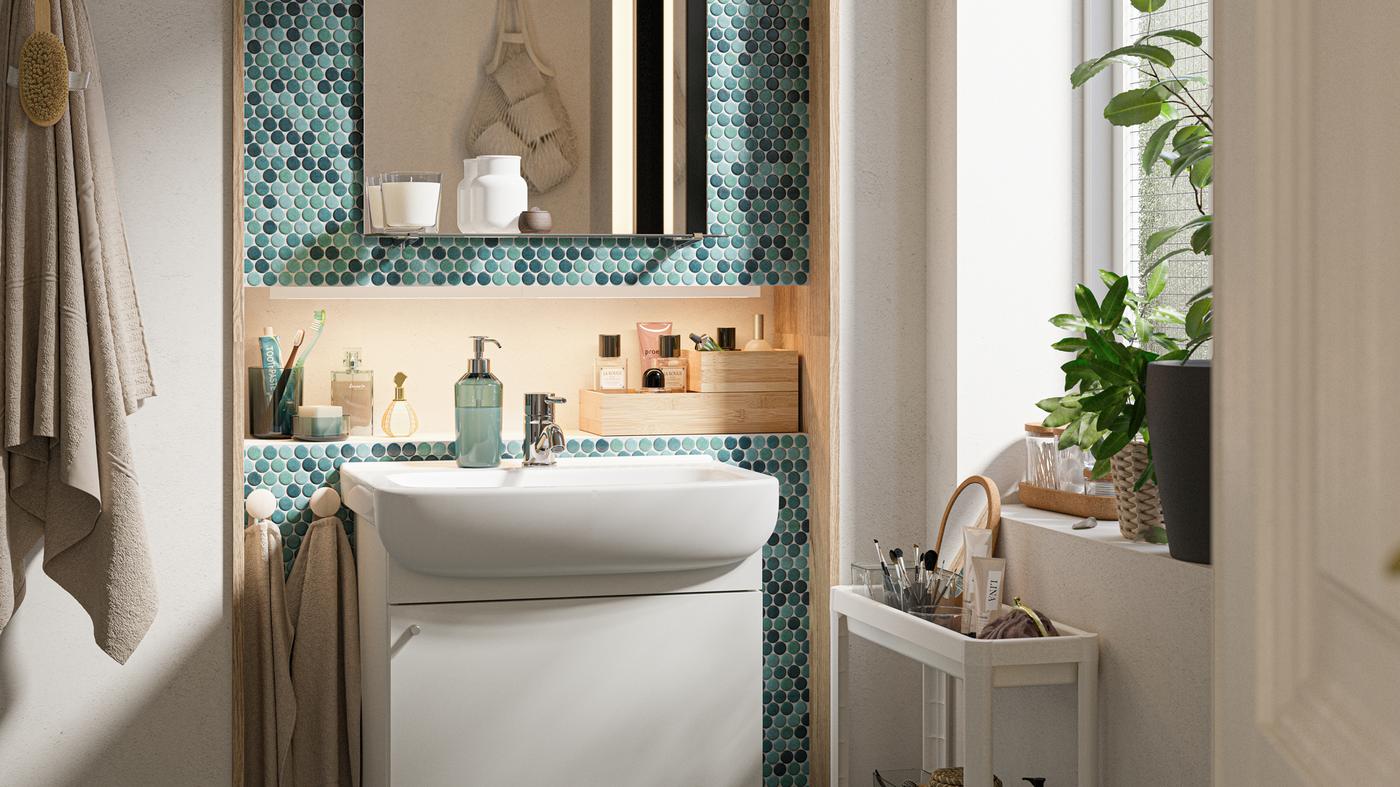 Un baño con azulejos verdes con un mueble de lavabo blanco, un espejo con estante y un carrito blanco con artículos de tocador en él.