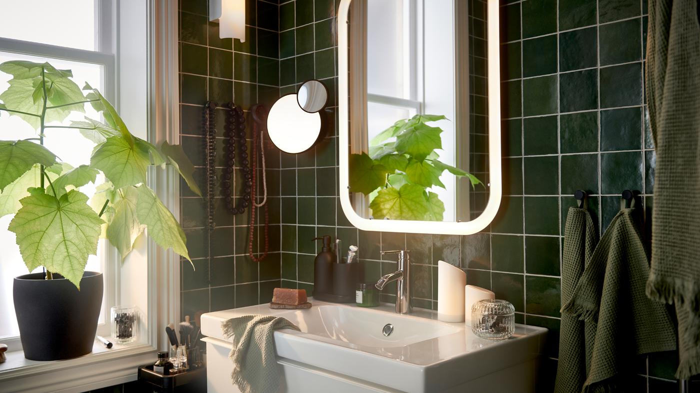 Un baño con espejo blanco con iluminación integrada sobre lavabo, además de toallas de invitados de color verde claro y una maceta con una planta.