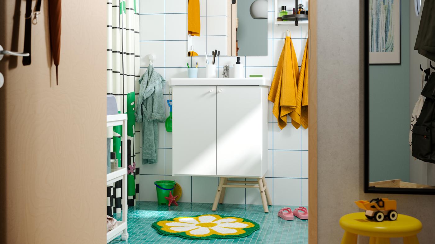 Un baño familiar con piso verde, toallas amarillas, tapete floral, ganchos de pared en diferentes alturas y muebles ENHET blancos.