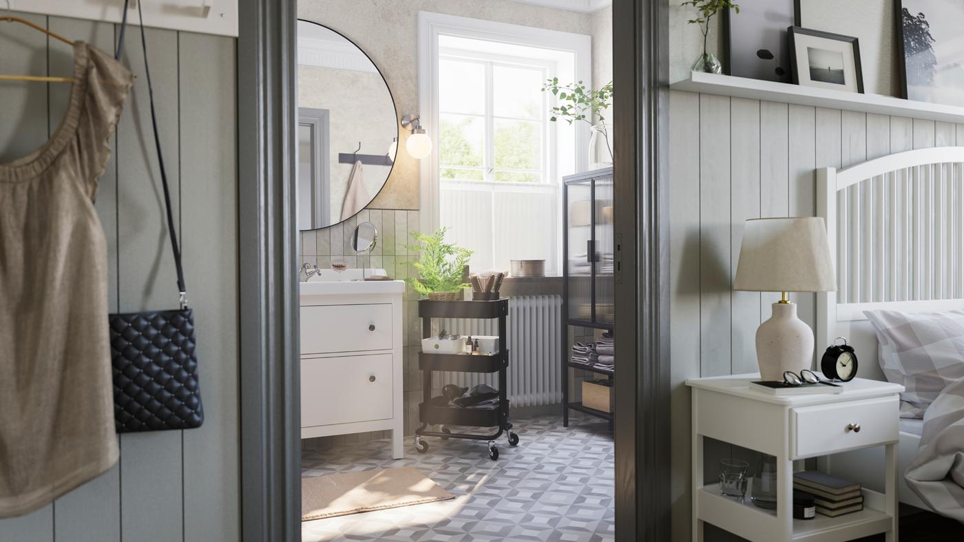 Un baño gris con una gran ventana tiene un mueble blanco HEMNES con lavabo, un carrito de negro RÅSKOG y un gabinete con puerta de cristal MOSSJÖN.