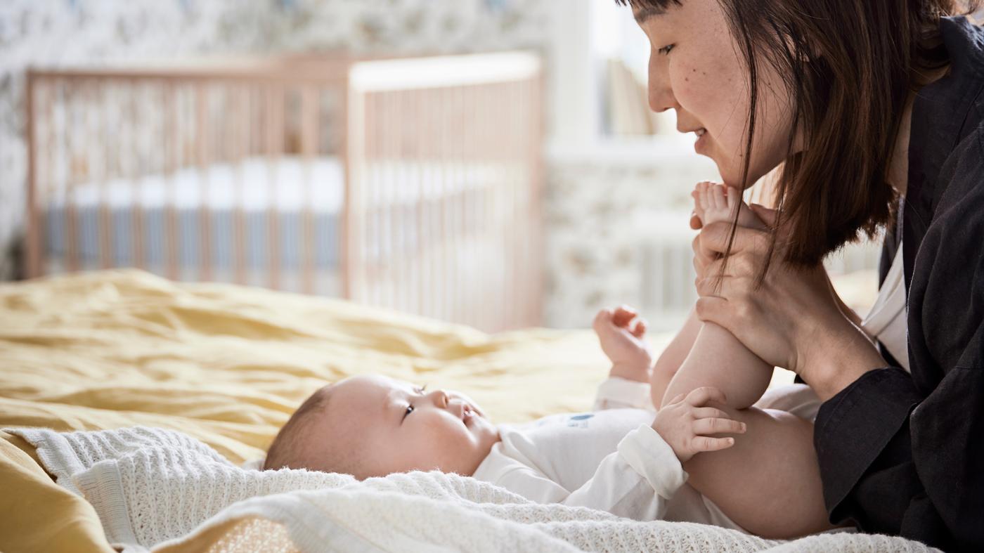 Un bébé à l’air heureux est couché sur le dos sur une couverture GULSPARV blanche étendue sur un lit. Sa maman lui tient les pieds et lui sourit.