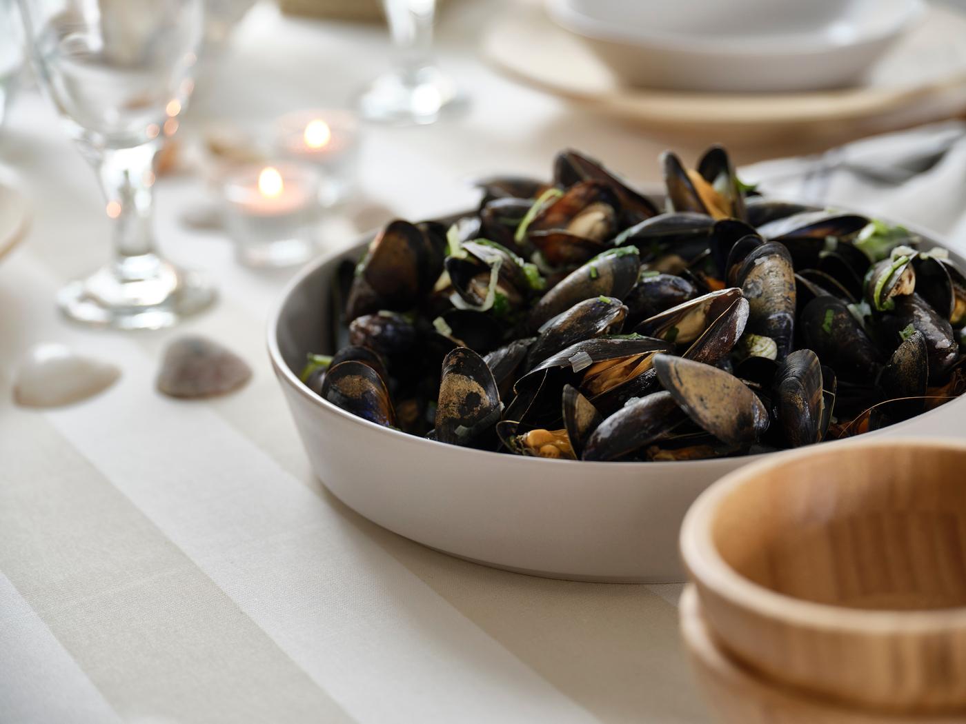 Un bol de moules posé sur une table dressée avec délicatesse, avec des verres, des bougies chauffe-plat et un saladier en bois à proximité.
