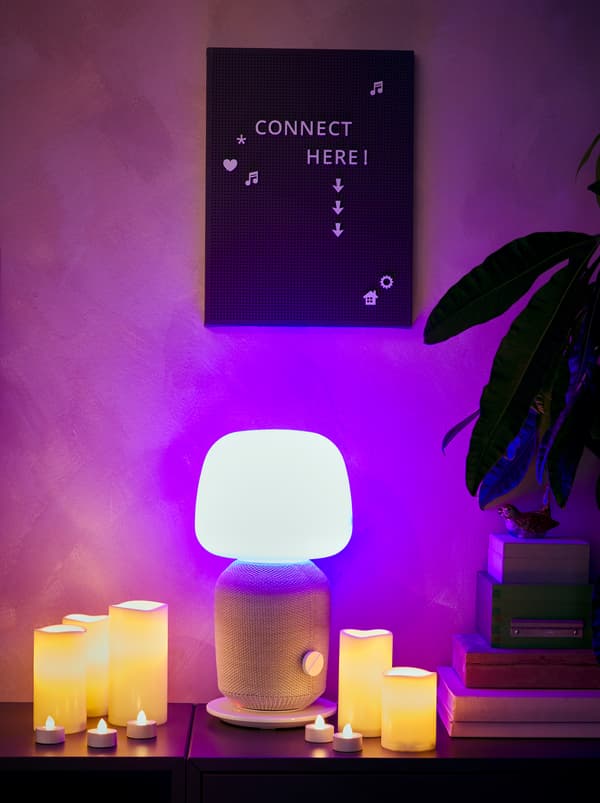 Un buffet avec une lampe SYMFONISK avec enceinte wifi intégrée, allumée et entourée de bougies blocs et bougies chauffe-plat LED, le tout surmonté d’un panneau SVENSÅS.