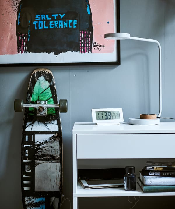 Un bureau blanc avec une tablette encastrée, au-dessus se trouve une lampe de bureau et une horloge digitale, un cadre avec un graffiti est suspendu au mur au-dessus d’un skateboard.