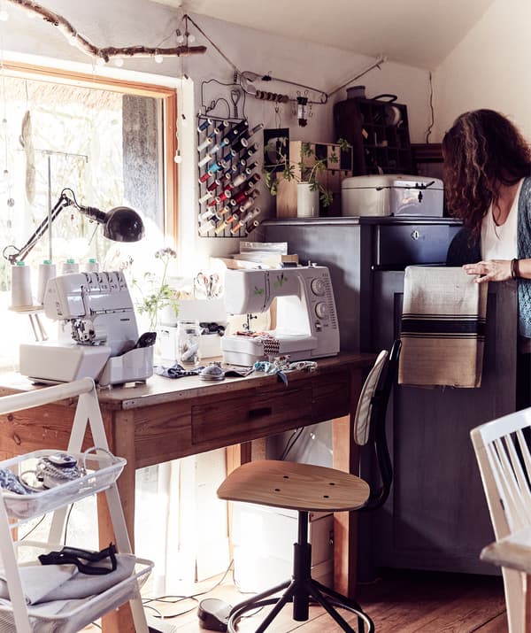 Un bureau en bois et une chaise pivotante devant une grande fenêtre et sur lequel sont posés des machines à coudre et du matériel artistique.