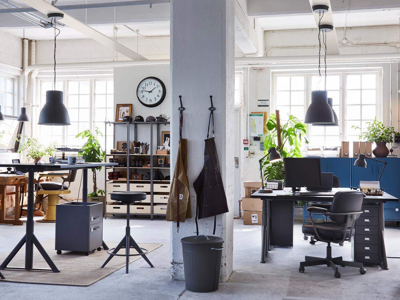 Un bureau industriel contenant des étagères, des espaces de travail avec bureaux assis-debout IDÅSEN et des tabliers suspendus à un pilier en béton.