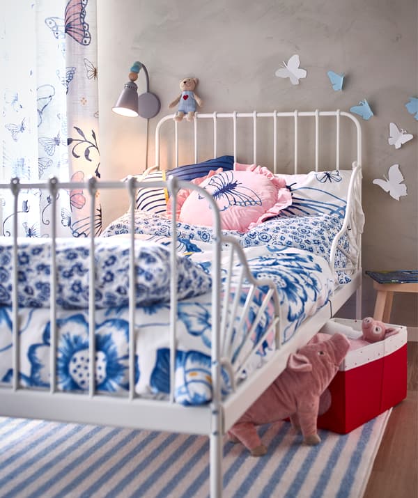 Un cadre de lit bleu recouvert d'un linge de lit floral, de coussins et de rideaux avec un motif de papillon, et des jouets sous le lit.