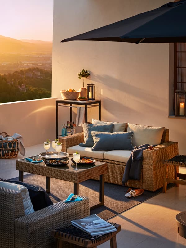 Un canapé d’extérieur avec des coussins blancs sur une terrasse avec un parasol, une table basse avec des rafraîchissements et un fauteuil d’extérieur.