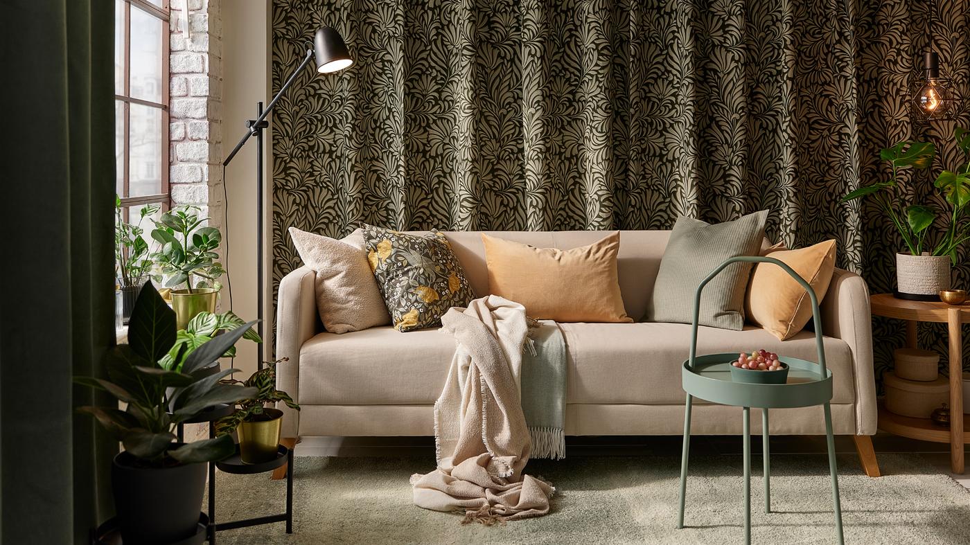 Un canapé LINANÄS beige avec plusieurs coussins sur un tapis gris-vert clair, devant des rideaux à motifs de feuilles accrochés à un mur.