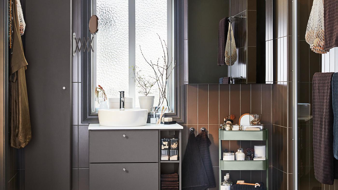 Un cuarto de baño con muebles de color gris oscuro, un armario con puerta de espejo y un carrito de color verde grisáceo.